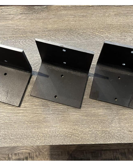 Three black steel L-shaped shelf brackets by Industrial Farm Co with mounting holes, displayed on weathered wood surface s...