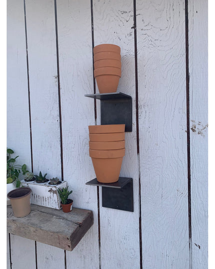 Black steel L-shaped shelf brackets mounted on white wooden wall holding stacks of terracotta pots, with succulent plants ...