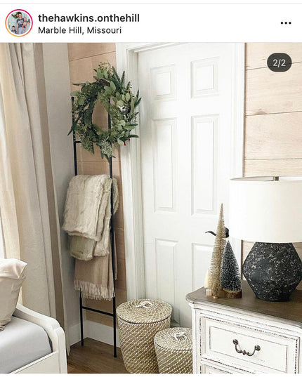 Black metal ladder with green wreath and cream throw blanket in farmhouse bedroom corner next to white dresser and wicker ...