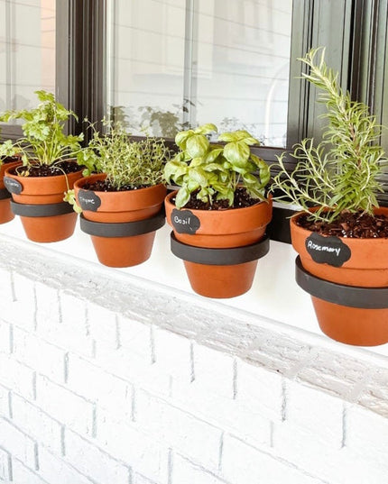 Terra cotta herb pots with black chalkboard labels displaying thyme, basil, and rosemary lined on white brick windowsill