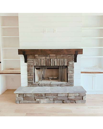 Rustic wooden fireplace mantel with black metal brackets installed above stone fireplace surround with built-in white shel...