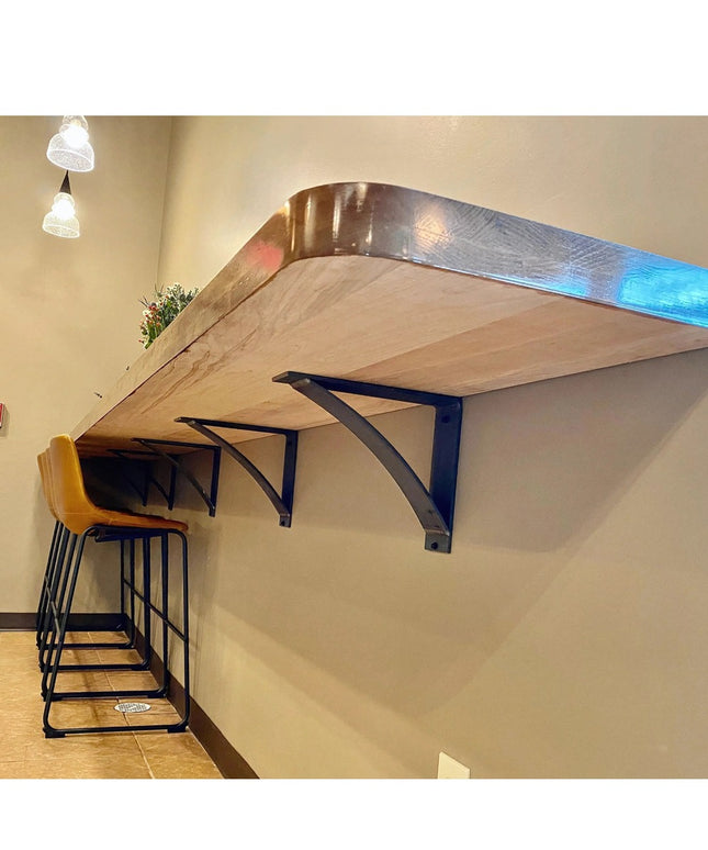 Black Hattie Style curved metal bracket supports holding floating wood shelf with yellow bar stools underneath by Industri...
