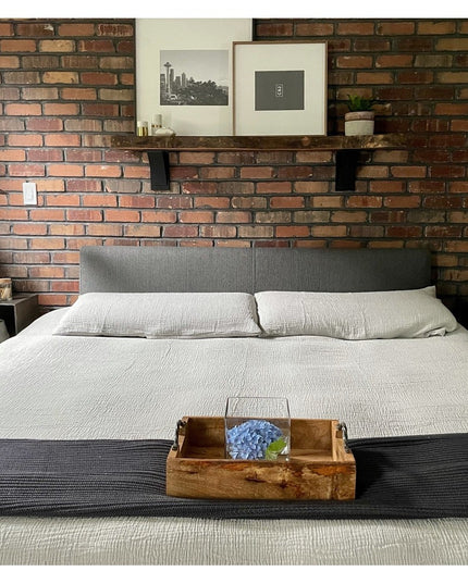 Industrial black steel shelf brackets supporting floating wood shelf above gray upholstered bed against exposed brick wall...