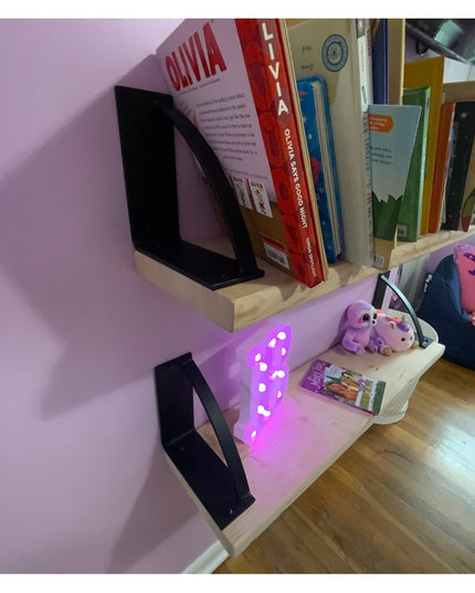 Black triangular Brie Bookshelf Brackets from Industrial Farm Co supporting floating wood shelves with books and purple LE...