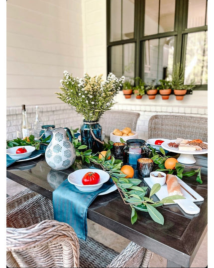 Modern outdoor dining table with blue table runner, fresh greenery garland, white plates with red tomatoes, and wicker cha...