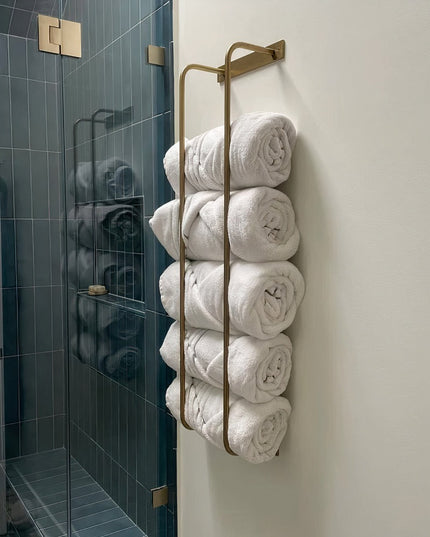 Brass wall-mounted Tyler Bathroom Towel Rack by Industrial Farm Co holding rolled white towels next to modern glass shower...