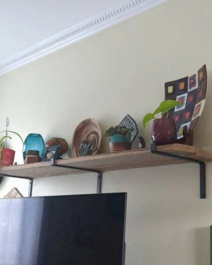 Black metal Z-style shelf brackets by Industrial Farm Co supporting wooden floating shelf with plants and decor above TV i...