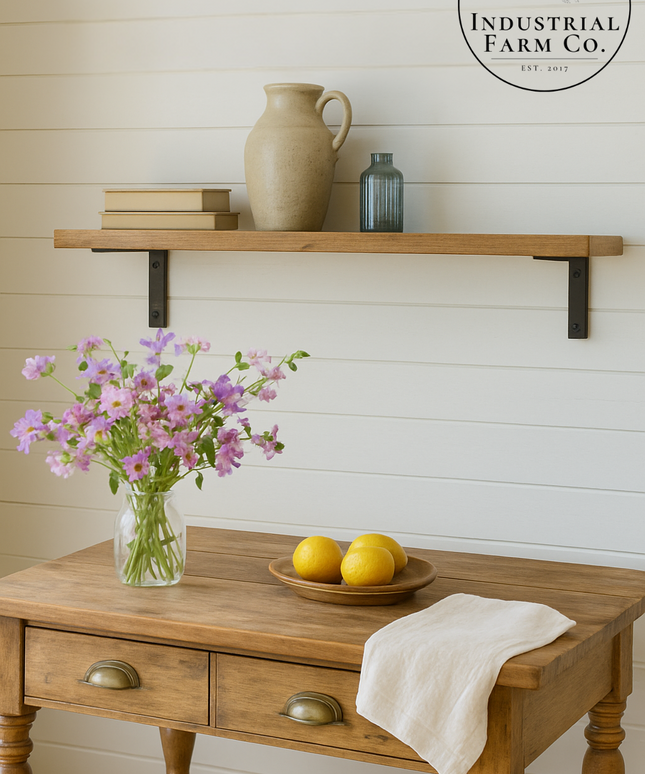 Wooden table with a vase of flowers, lemons, and a towel, with a shelf above featuring a pitcher and bottles against a white paneled wall.