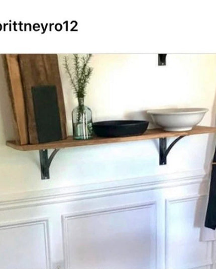 Black steel industrial shelf brackets supporting wooden shelf with decorative bowls and vase above white wainscoting